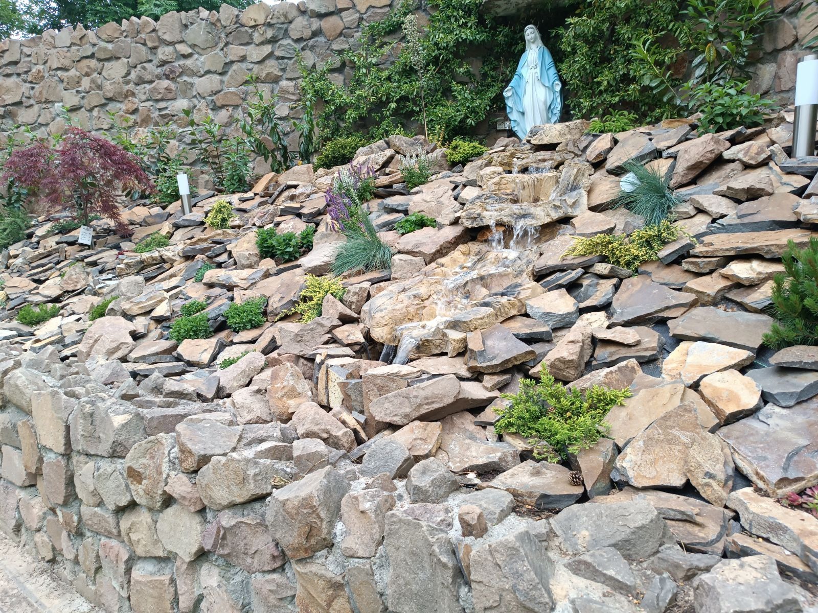 Kaskada wodna z kamieni w ogrodzie, z figurką Matki Boskiej na szczycie, otoczona różnorodną roślinnością.