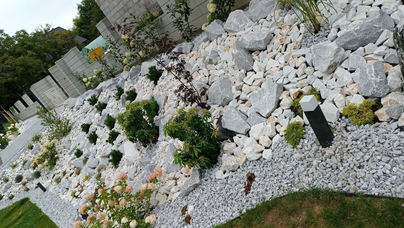 Aranżacja skalniaka w Libertowie z wykorzystaniem białych i szarych kamieni różnej wielkości, niskich krzewów ozdobnych i lampy ogrodowej.