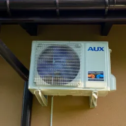 Biała jednostka zewnętrzna klimatyzacji marki AUX zamontowana na ścianie budynku o brązowej elewacji, widoczny fragment rynny i niebieskie niebo z drzewami w tle.