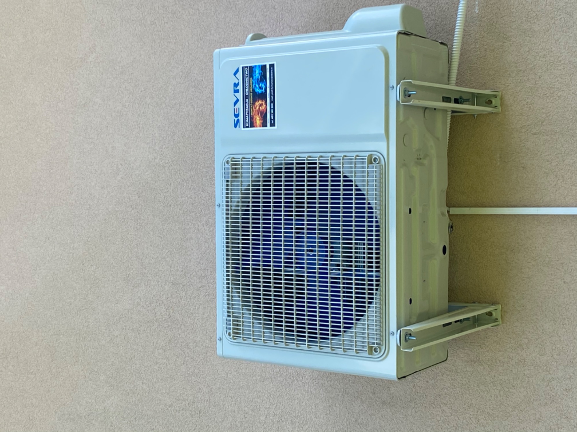 Biała jednostka zewnętrzna klimatyzatora zamontowana na ścianie o szorstkiej fakturze, widoczna etykieta producenta SEVRA z informacjami kontaktowymi.