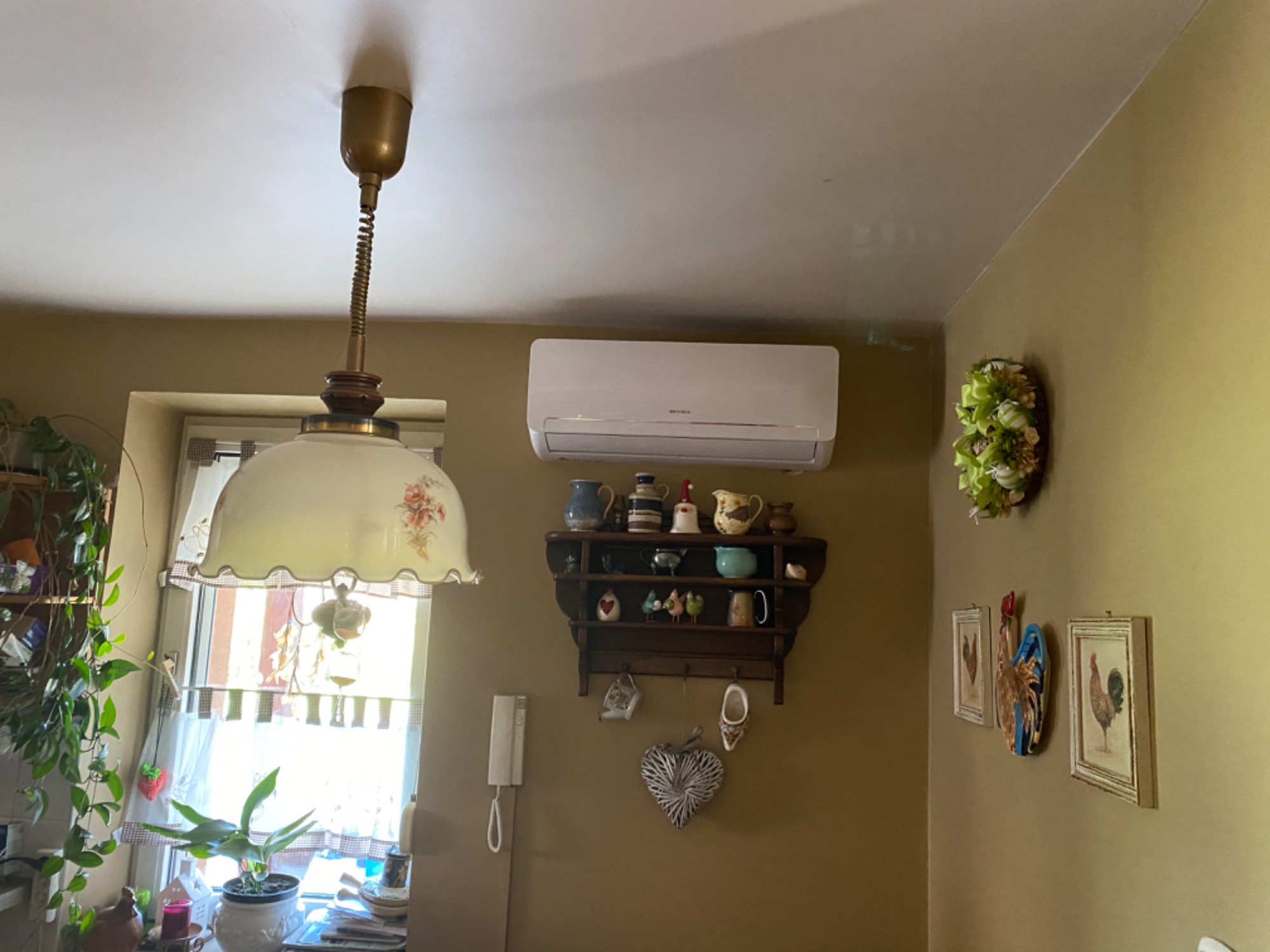 Wnętrze pokoju z zamontowaną klimatyzacją na ścianie nad półką z dekoracjami, widoczna lampa wisząca w stylu retro.