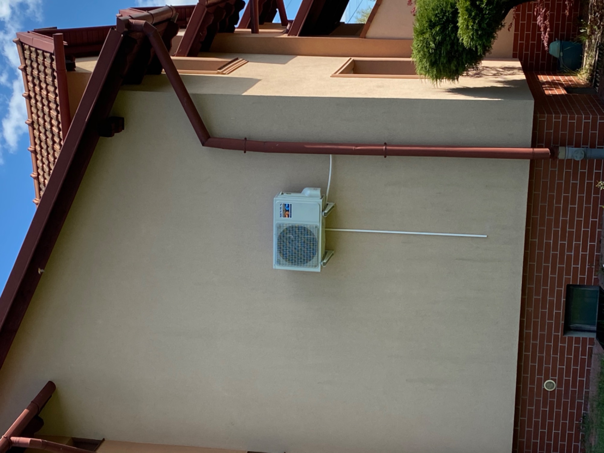 Zewnętrzna jednostka klimatyzacji zamontowana na elewacji domu jednorodzinnego, widoczne rury odprowadzające, brązowy dach i rynna.