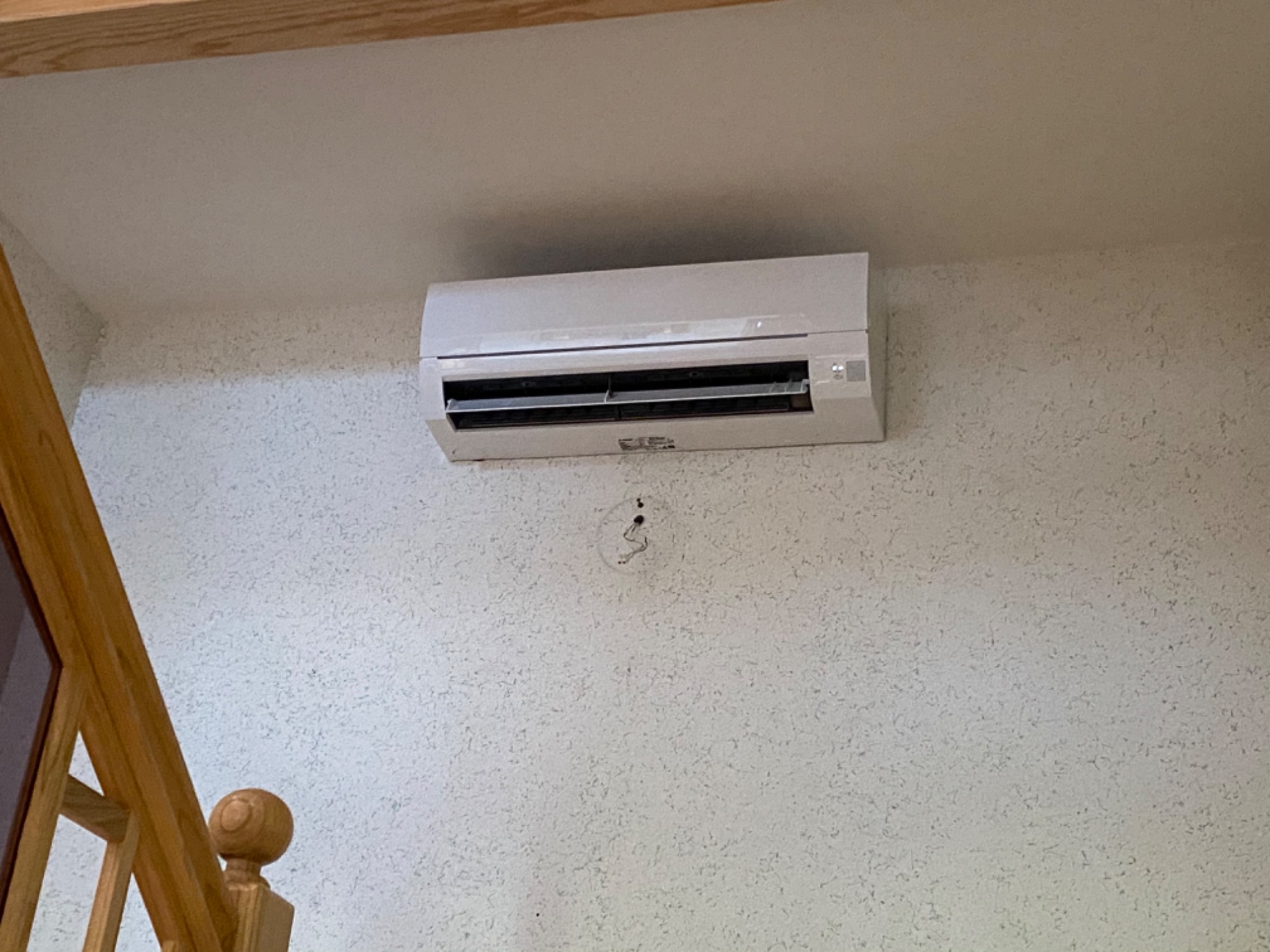 Biała jednostka klimatyzacji ściennej zamontowana na teksturowanej ścianie wewnątrz pomieszczenia, widoczne otwory po przewodach, obok fragment drewnianej balustrady schodów.