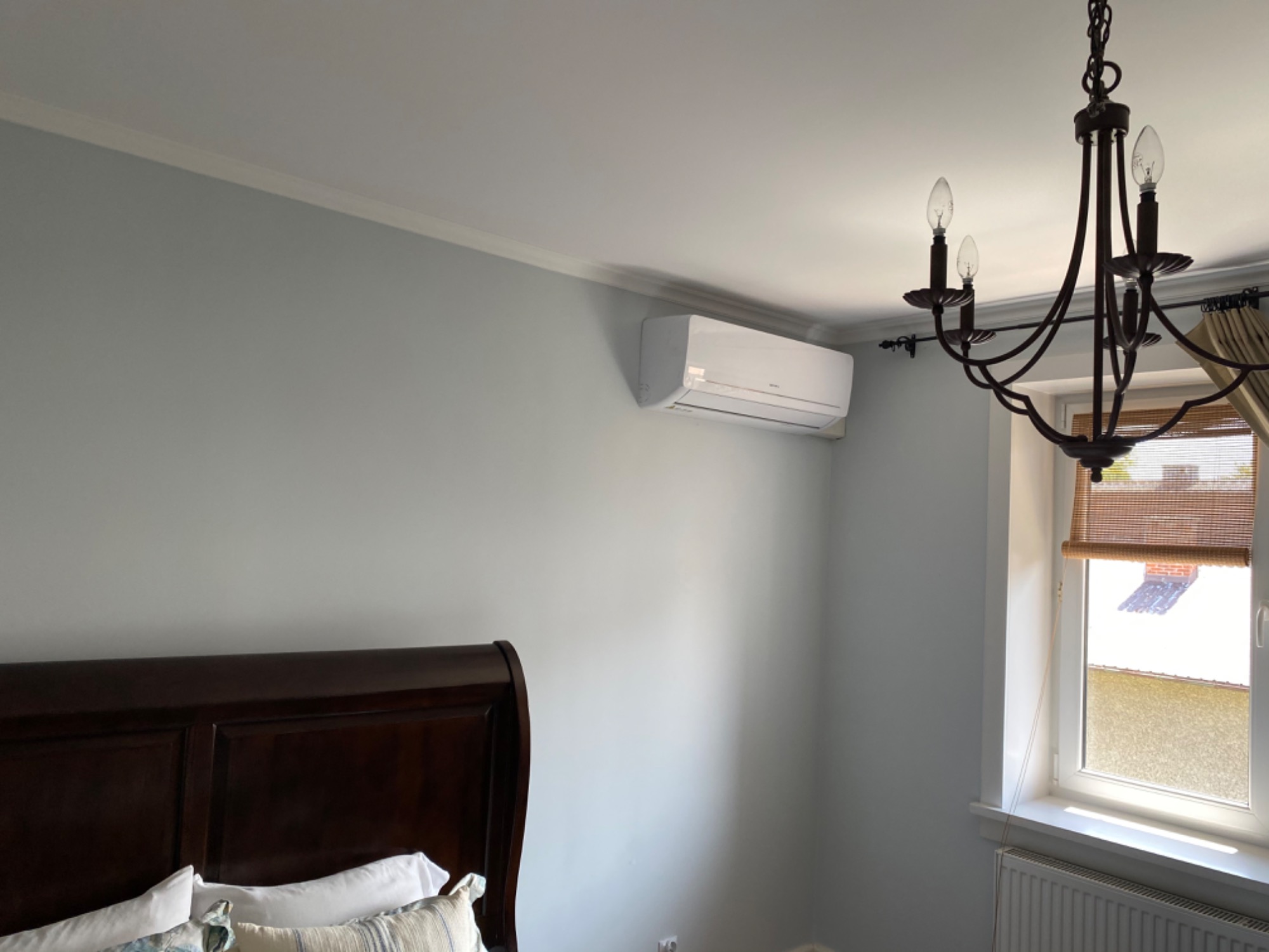 Biały klimatyzator ścienny zamontowany w sypialni z szarą ścianą, widoczny fragment ciemnego, drewnianego łóżka, okno z bambusową roletą i żyrandol w stylu retro.