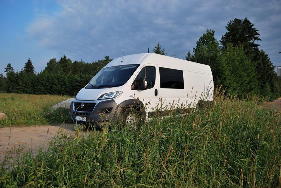 Biały furgon z przyciemnianymi szybami, zaparkowany na polnej drodze obok wysokiej trawy, na tle lasu i pochmurnego nieba.