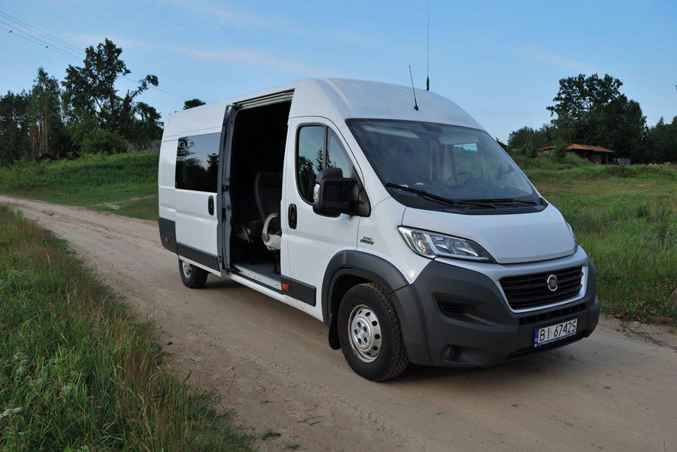 Biały bus z otwartymi drzwiami bocznymi stoi na polnej drodze, widoczne wnętrze z czarnymi fotelami i kierownicą.