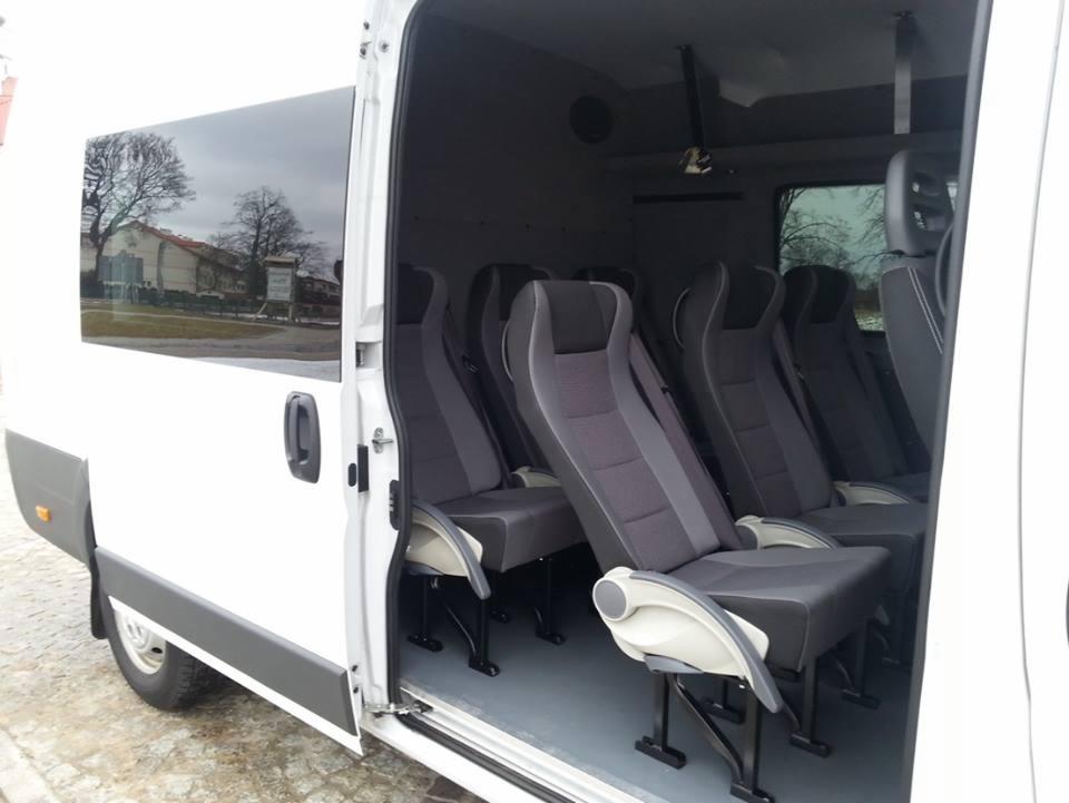 Wnętrze białego busa z szarymi fotelami pasażerskimi, widok z otwartych drzwi bocznych na rząd foteli z wysokimi zagłówkami i podłokietnikami, w tle widoczny budynek odbijający się w szybie.