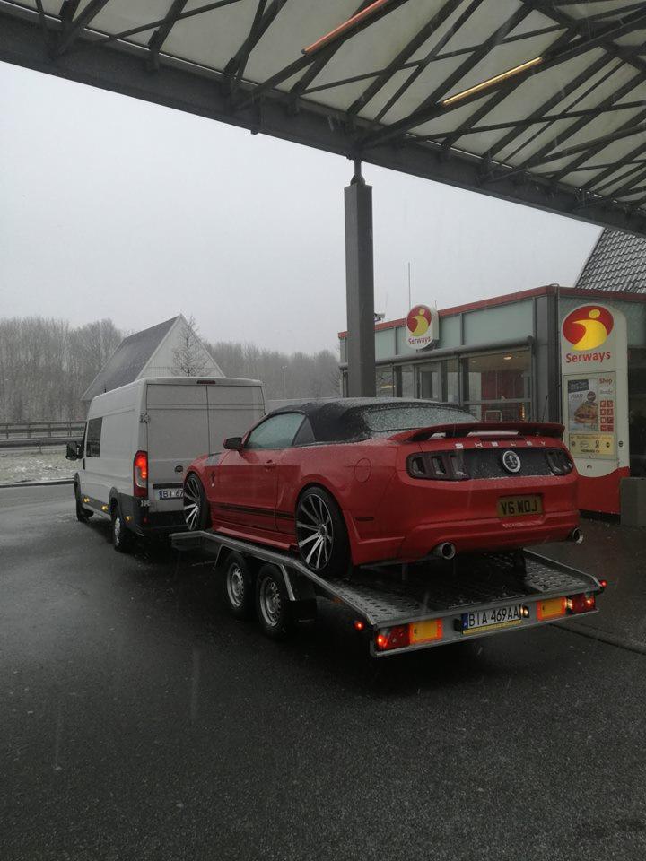 Czerwony sportowy kabriolet Ford Mustang V6 na niskopodwoziowej przyczepie, ciągniętej przez białego busa na stacji benzynowej w pochmurny dzień.