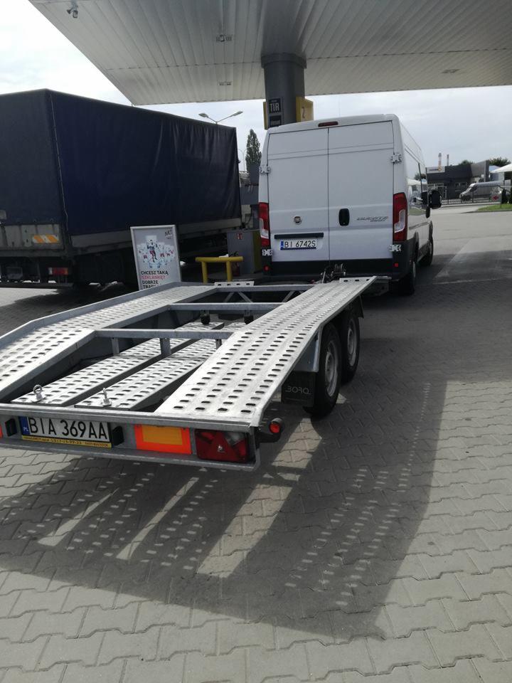 Laweta z podłączonym białym busem na stacji benzynowej, w tle widoczna naczepa ciężarówki z plandeką.