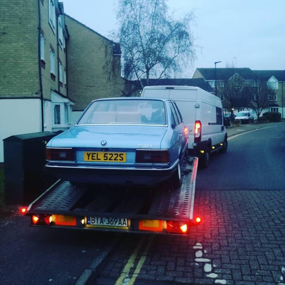 Błękitny, klasyczny samochód BMW 518 z żółtymi tablicami rejestracyjnymi YEL 522S, transportowany na platformie lawety holowanej przez białego busa, w tle zabudowa miejska.