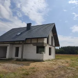 Niedokończony, murowany dom jednorodzinny z ciemną dachówką i kominem, widoczny w stanie surowym otwartym na tle błękitnego nieba z chmurami i zalesionego terenu.