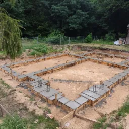 Zalane betonem szalunki pod fundamenty domu na piaszczystym terenie, widoczne wystające pręty zbrojeniowe, w tle zalesiony teren i budynek gospodarczy.