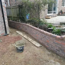 Nowo wymurowana, zakrzywiona ściana oporowa z czerwonej cegły w ogrodzie, widoczny kubeł i deska szalunkowa, w tle dom z jasnej cegły.