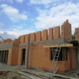 Budowa domu jednorodzinnego z czerwonej cegły, widoczne ściany w trakcie murowania, stalowe pręty zbrojeniowe wystające ponad mury, drewniane podpory, drabina oparta o ścianę, błękitne niebo...