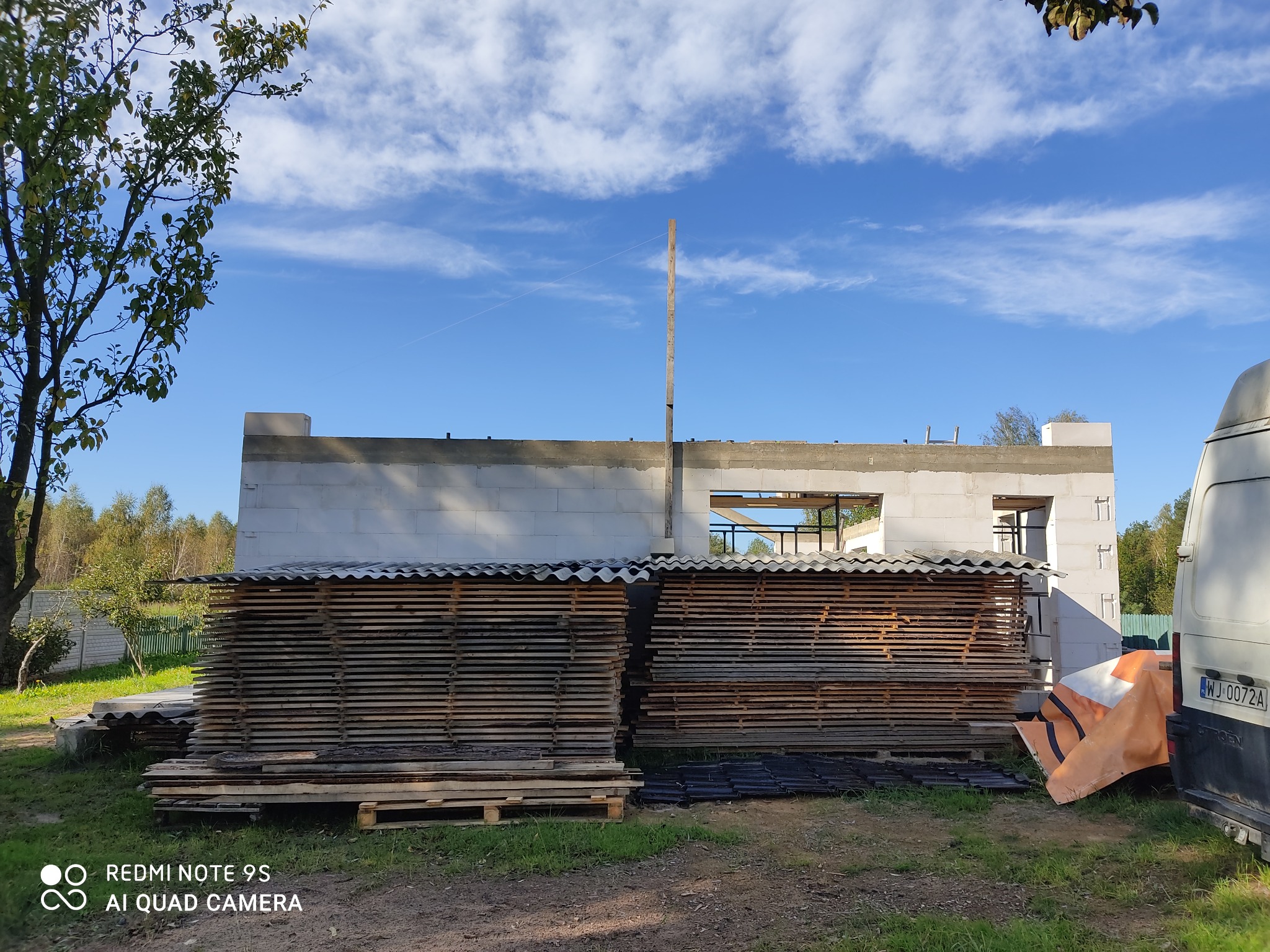 Wznoszony budynek z betonu komórkowego, widoczne przygotowane deski pod konstrukcję dachu, na pierwszym planie składowane płyty, w tle błękitne niebo z białymi chmurami.