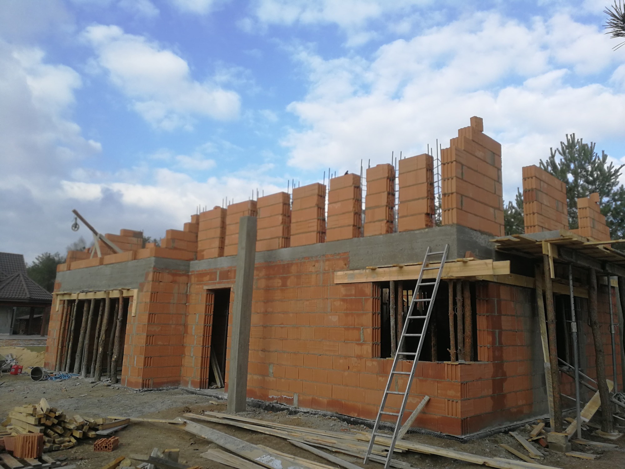 Budowa domu jednorodzinnego z czerwonej cegły, widoczne ściany w trakcie murowania, stalowe pręty zbrojeniowe wystające ponad mury, drewniane podpory, drabina oparta o ścianę, błękitne niebo...