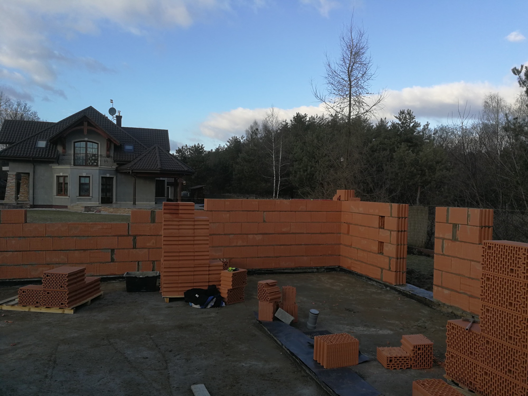 Budowa domu jednorodzinnego: wznoszone ściany z czerwonej ceramiki poryzowanej, widoczny fragment domu w tle i las.