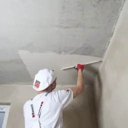 Pracownik w czerwonych rękawicach nakłada gładź szpachlową na surowy betonowy sufit za pomocą długiej szpachelki, widoczna ściana po prawej stronie, na ubraniu i czapce logo firmy budowlanej.