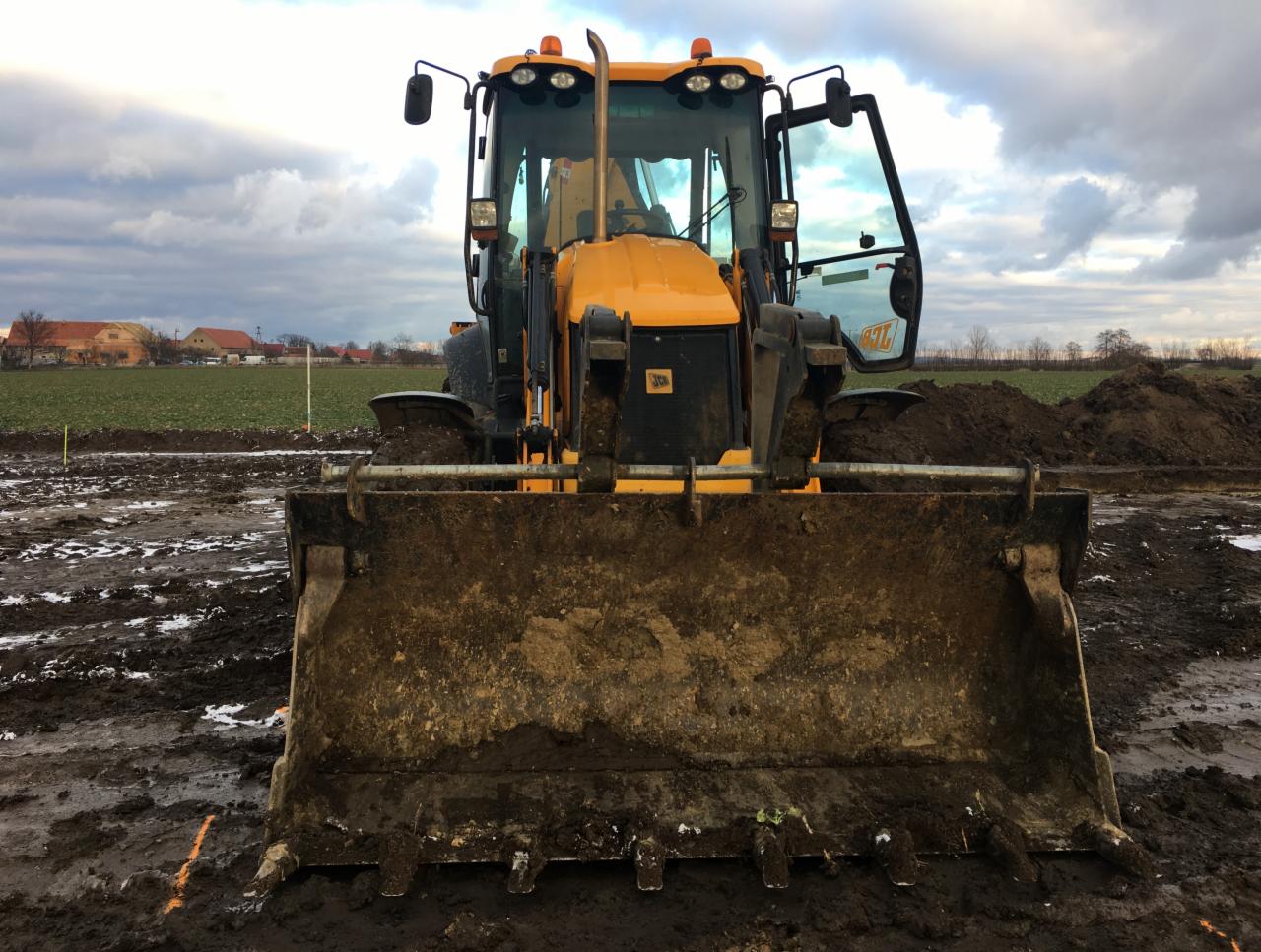 Żółta koparko-ładowarka JCB z otwartymi drzwiami kabiny, stojąca na błotnistym terenie z widocznymi śladami opon i pryzmami ziemi, w tle pole i zabudowania wiejskie pod pochmurnym niebem.