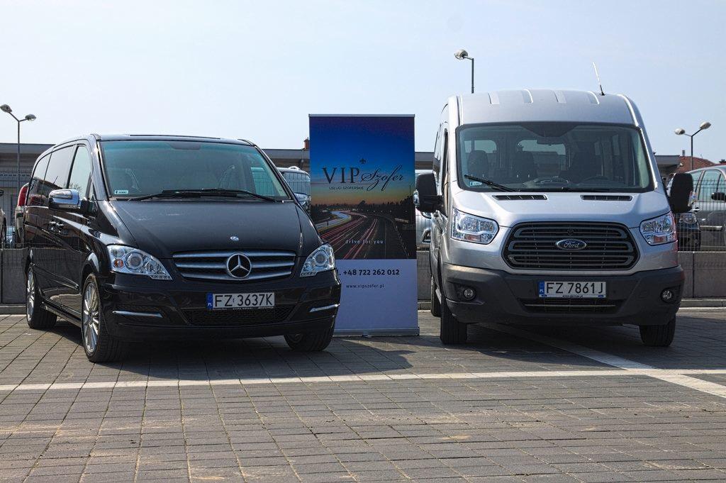 Dwa busy: czarny Mercedes i srebrny Ford, zaparkowane obok roll-upu reklamującego firmę transportową VIP Szofer z numerem telefonu i adresem strony internetowej.