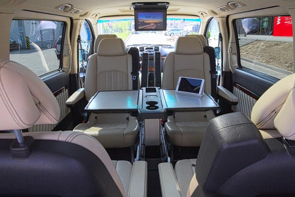 Wnętrze luksusowego busa z beżowymi skórzanymi fotelami, rozkładanymi stolikami, uchwytami na napoje i ekranem LCD podwieszonym pod sufitem, widok z tylnych siedzeń.