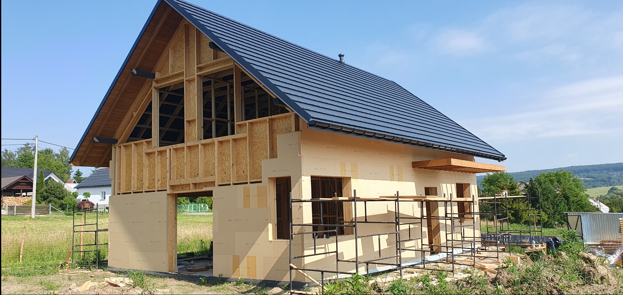Budowa domu w technologii szkieletowej: widok na konstrukcję z drewna i płyt OSB, czarny dach z dachówki ceramicznej, rusztowania przy elewacji.