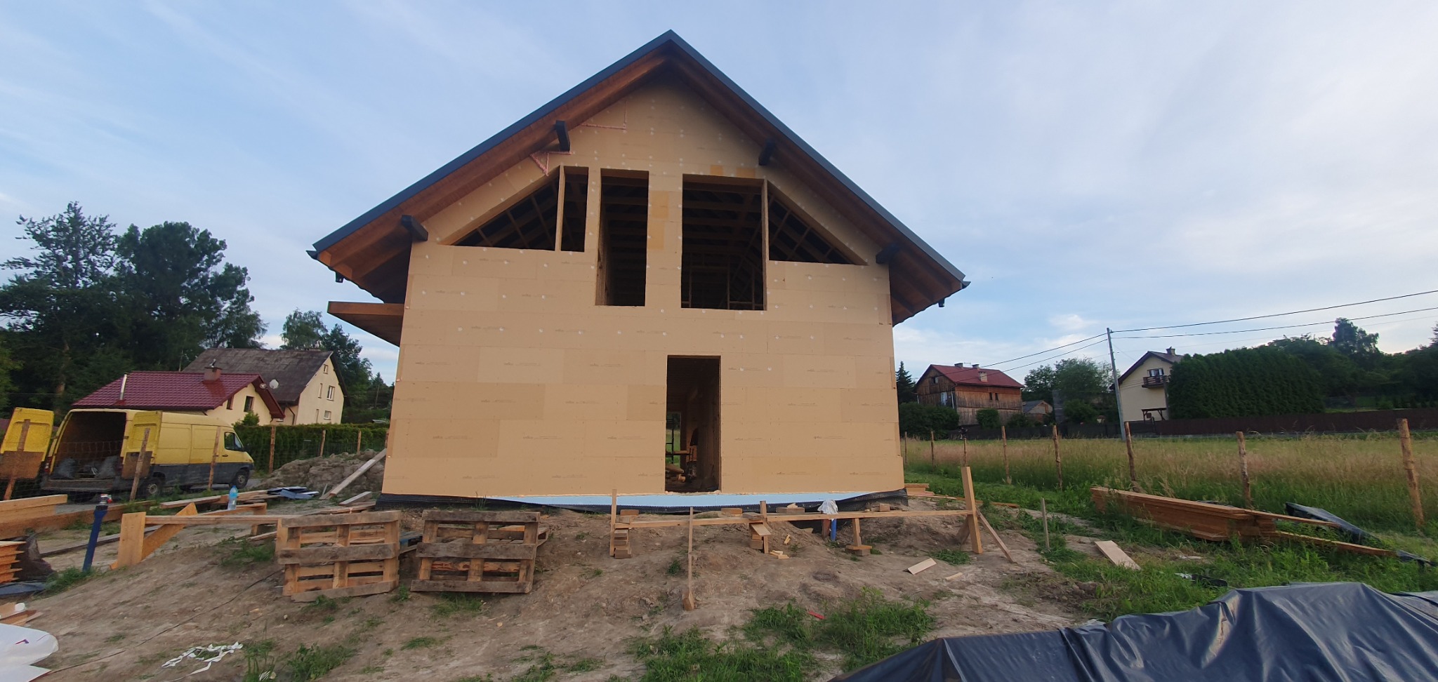 Surowy stan domu w konstrukcji szkieletowej z widoczną elewacją z płyt, drewnianą konstrukcją dachu, paletami i elementami rusztowania na nierównym terenie.