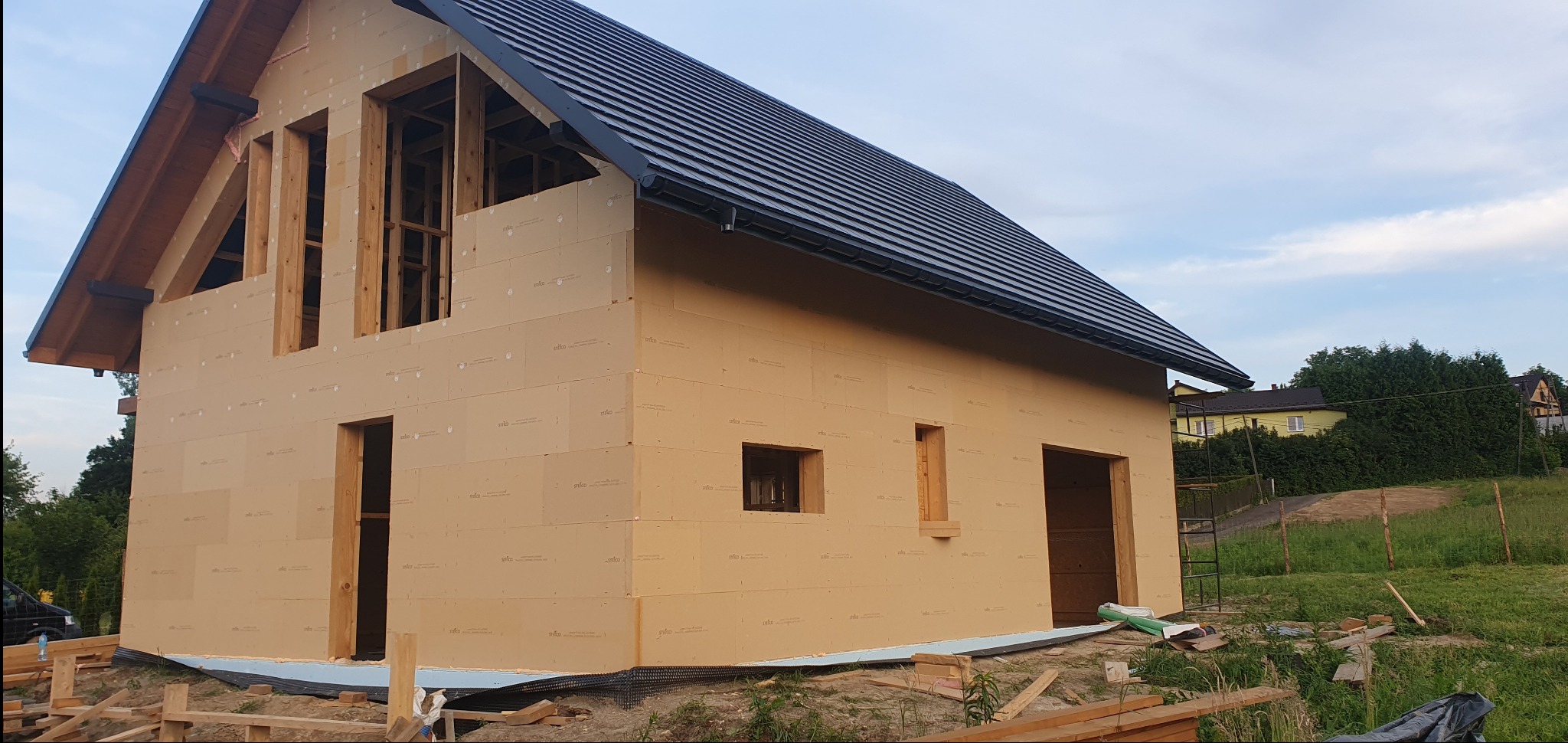 Surowy stan budynku z czarnym dachem, obłożonego jasnymi płytami izolacyjnymi z widocznymi otworami okiennymi i drzwiowymi, na tle zieleni i błękitnego nieba.