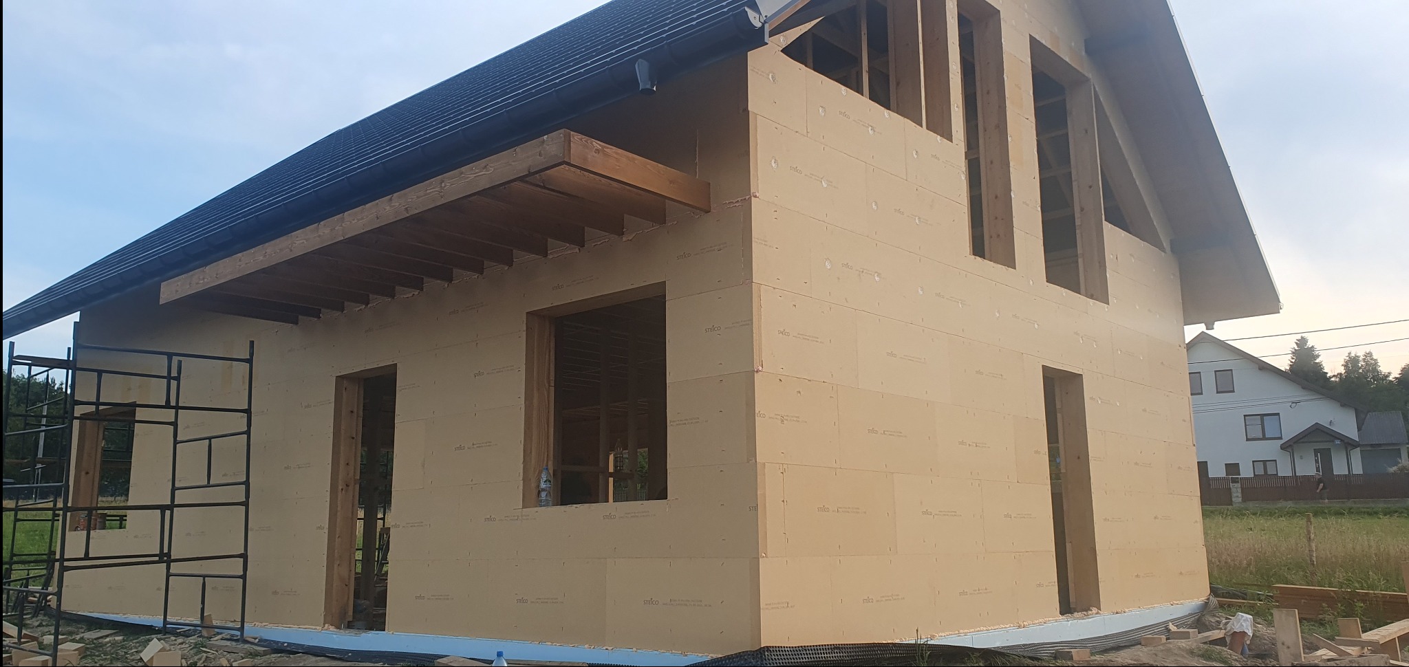 Surowy stan domu w konstrukcji szkieletowej z widocznym drewnianym szkieletem i płytami izolacyjnymi na elewacji bocznej, z rusztowaniem po lewej stronie i fragmentem sąsiedniego domu w tle.