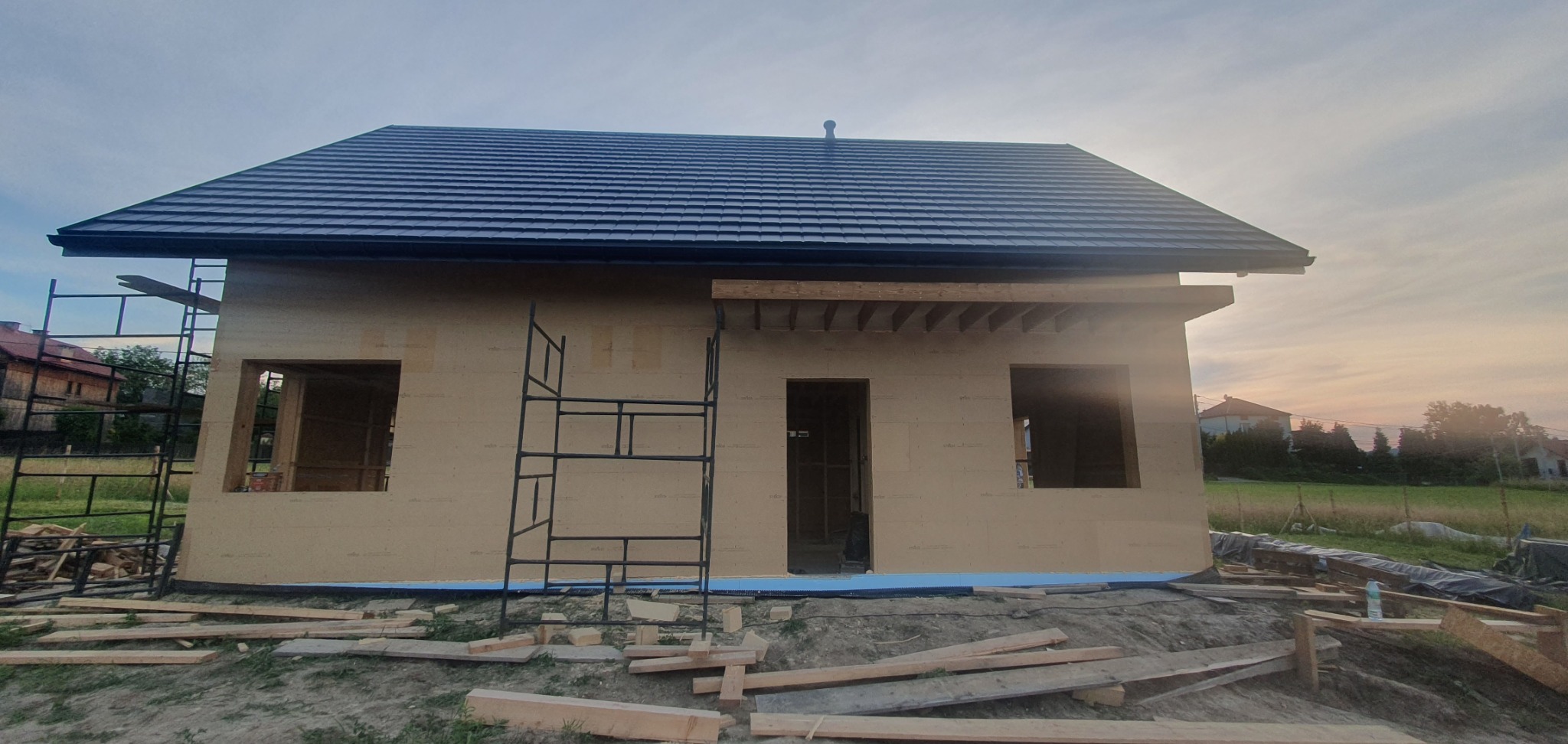 Surowy stan budynku z drewnianą konstrukcją szkieletową, widoczne rusztowanie i przygotowane deski, dach pokryty blachodachówką, tło stanowi zachodzące słońce.