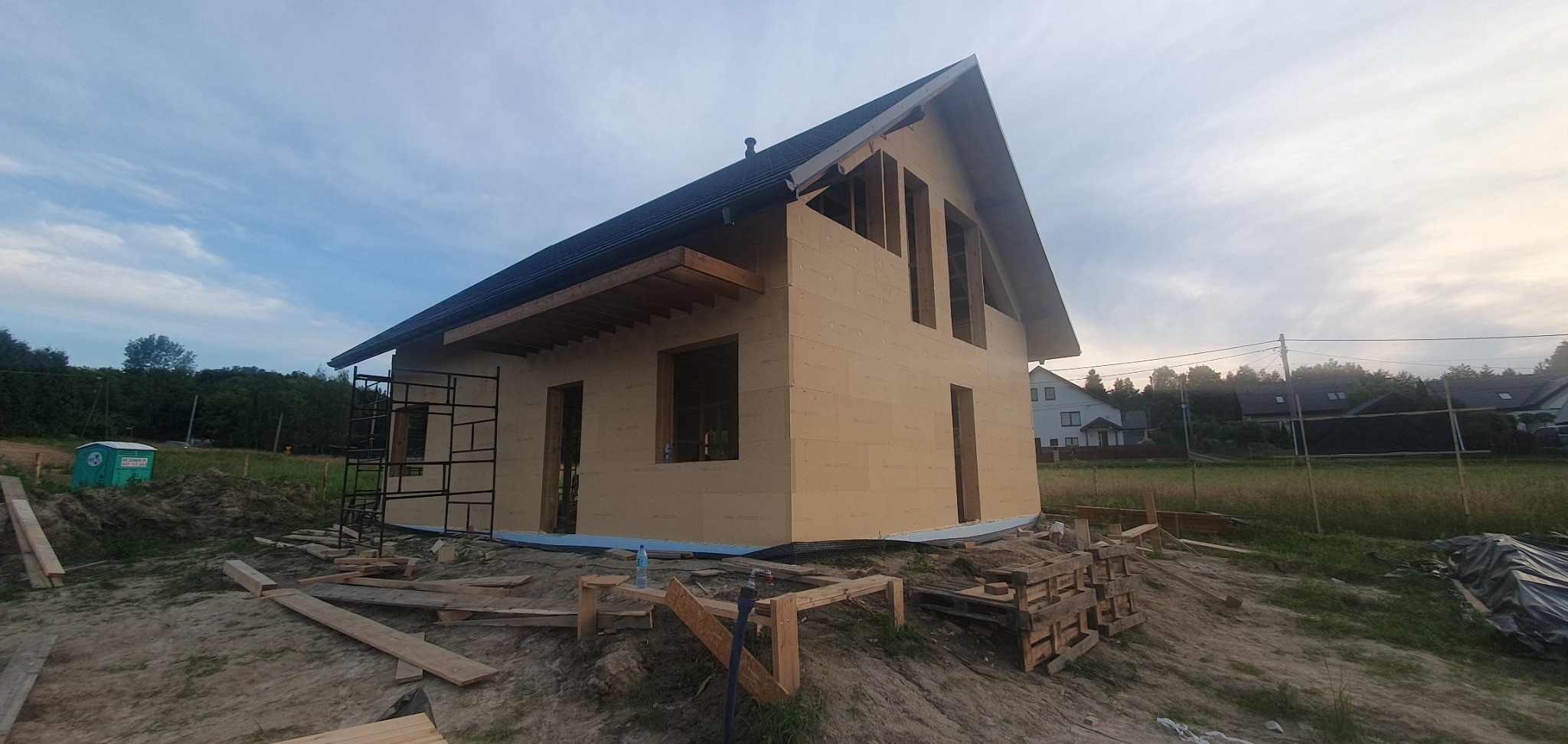 Surowy stan budynku w technologii szkieletowej z widocznymi rusztowaniami, drewnianymi elementami konstrukcyjnymi i przygotowanym podłożem pod elewację na tle zieleni i zabudowy jednorodzinnej.