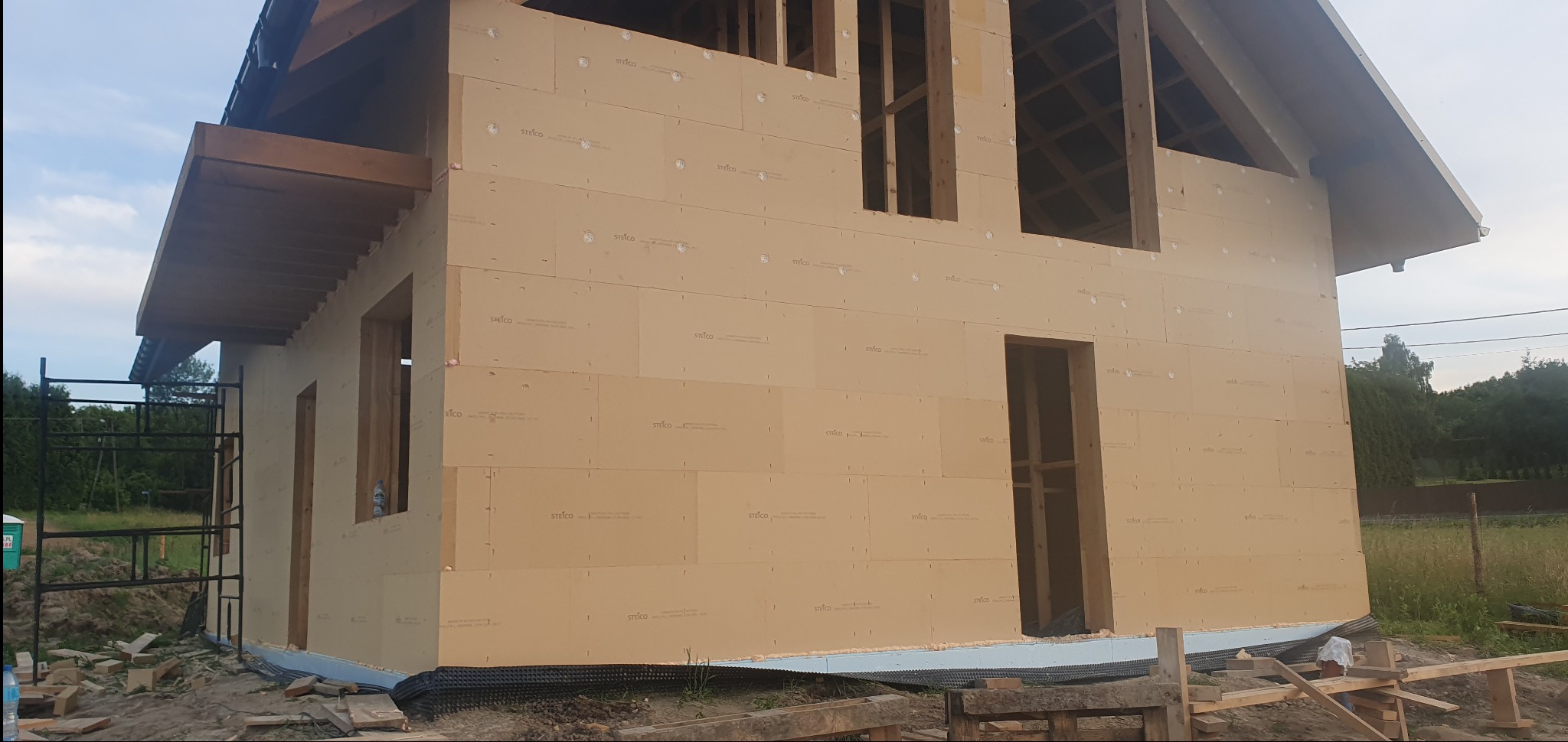 Surowy stan budynku z drewnianą konstrukcją i płytami izolacyjnymi STEICO, widoczne rusztowanie i przygotowany teren wokół.