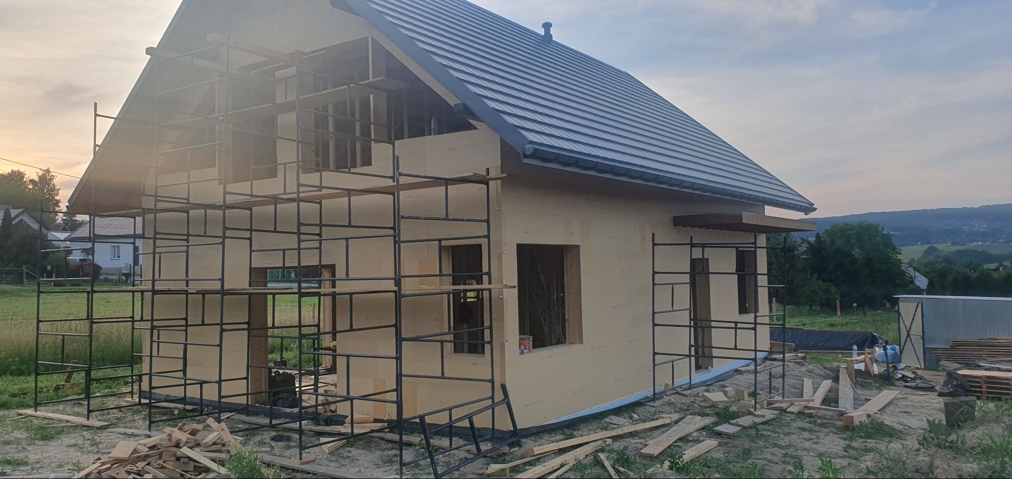 Surowy stan domu szkieletowego z ciemną dachówką, otoczonego rusztowaniem i drewnianymi odpadami budowlanymi na tle zachodzącego słońca.