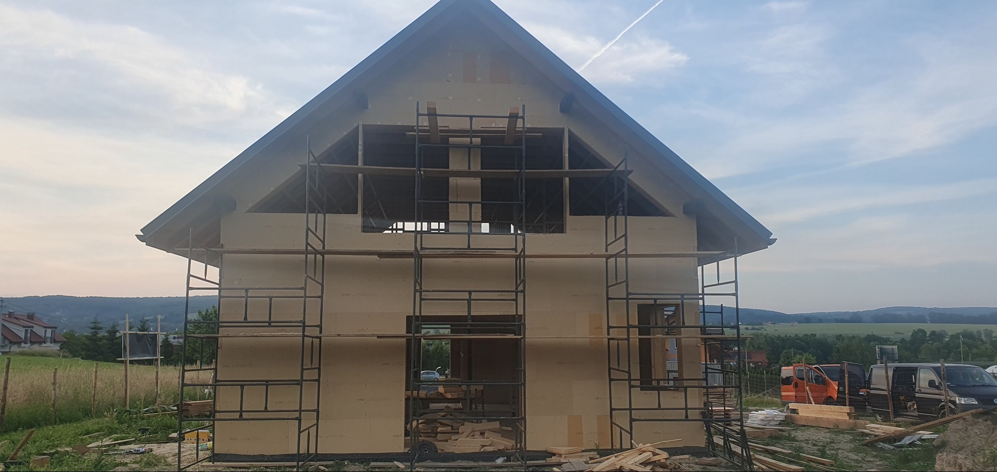 Surowy stan domu szkieletowego w trakcie budowy, z widocznymi rusztowaniami i przygotowanymi otworami okiennymi, na tle pagórkowatego krajobrazu.