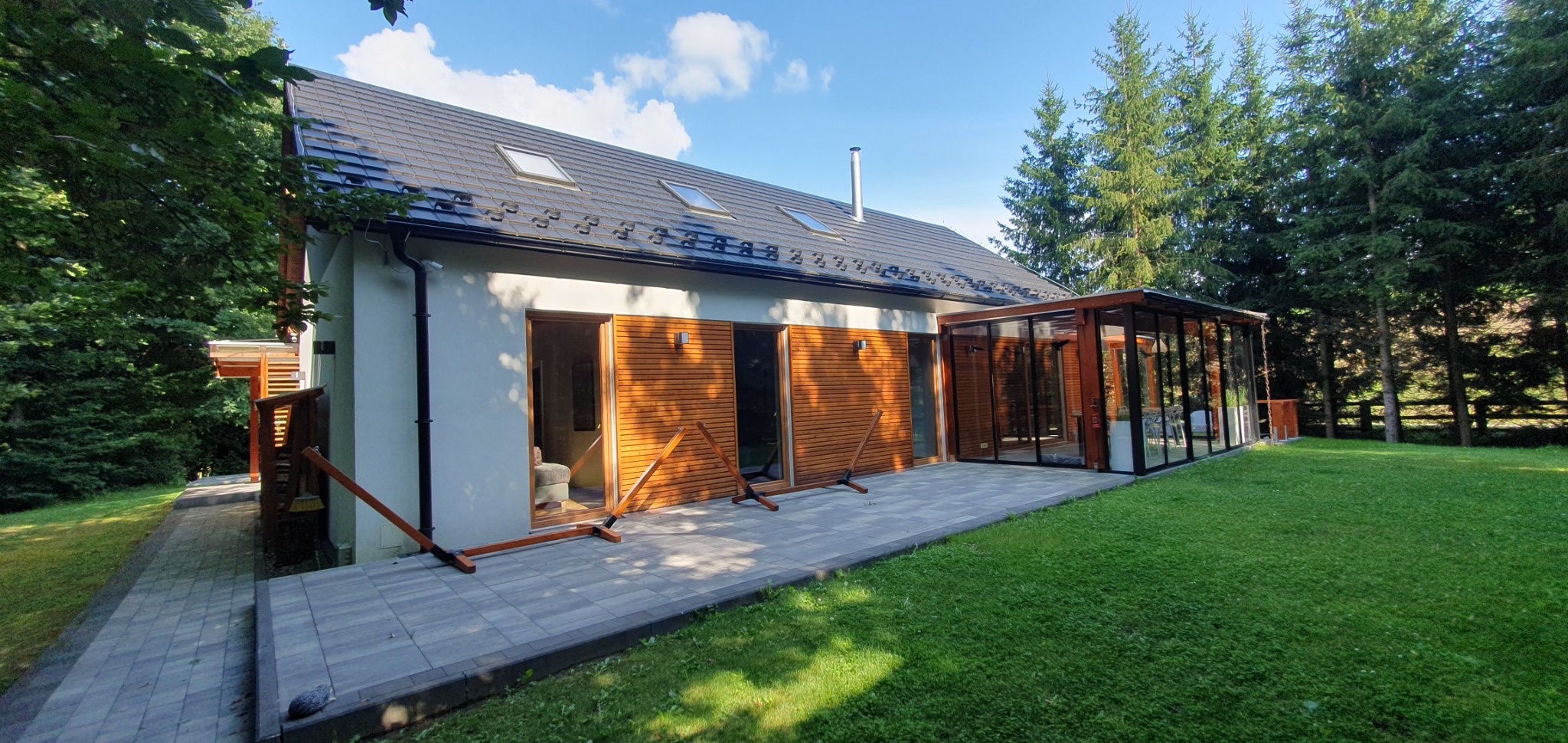 Nowoczesny dom z elewacją z jasnego tynku i drewnianą okładziną, przeszklona weranda, szary dach z oknami połaciowymi, zielony trawnik i drzewa w tle.