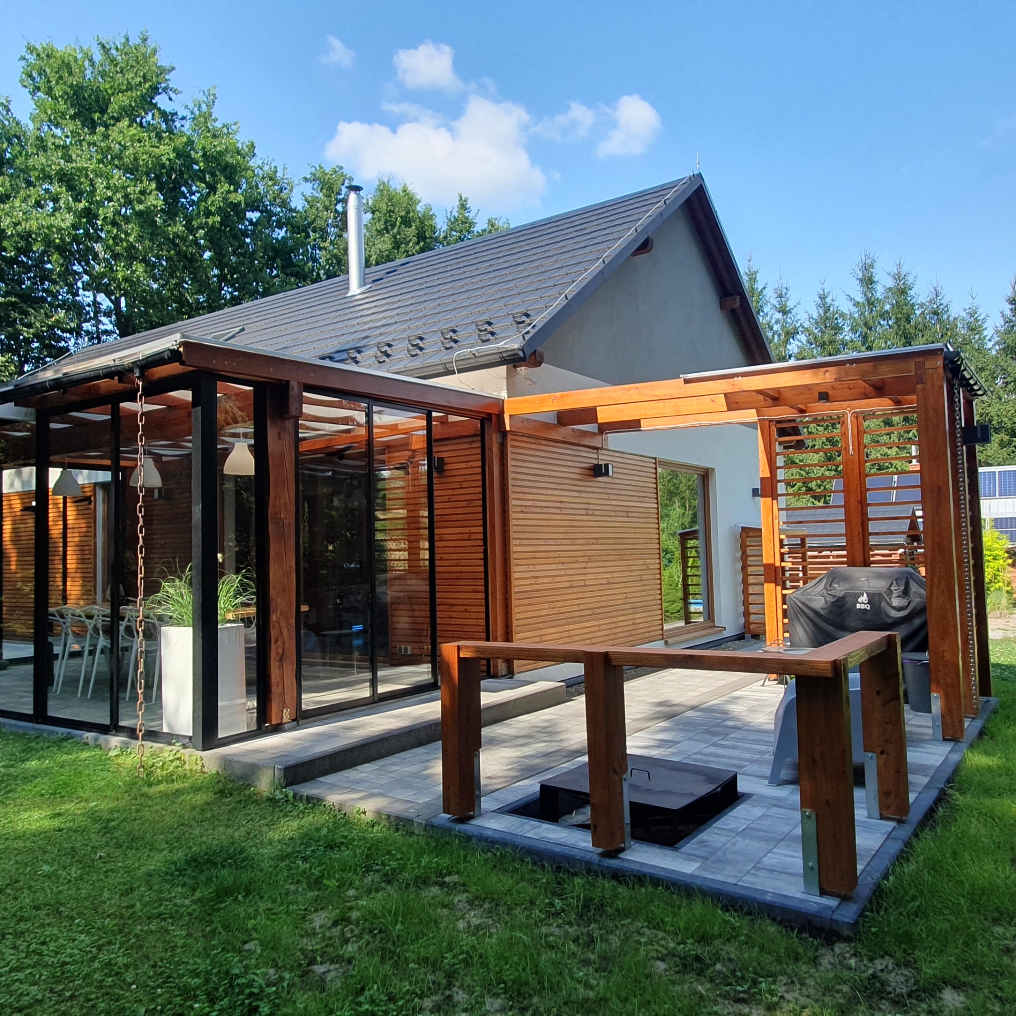 Nowoczesny dom z drewnianą elewacją, szklaną werandą i zadaszonym tarasem z grillem, otoczony zielenią.