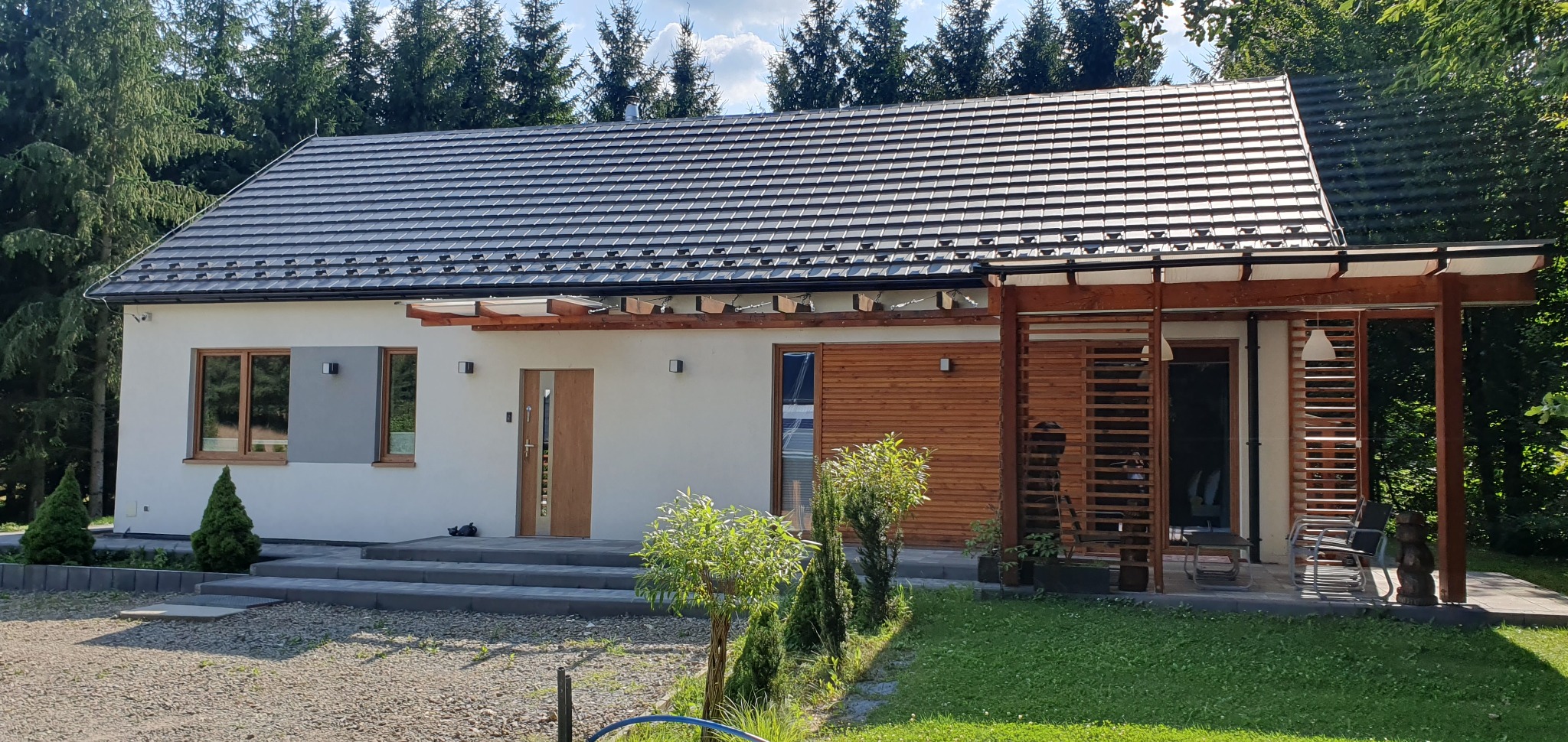 Gotowy dom szkieletowy z szarą dachówką, drewnianą pergolą i elementami wykończenia z naturalnego drewna, otoczony zielenią.