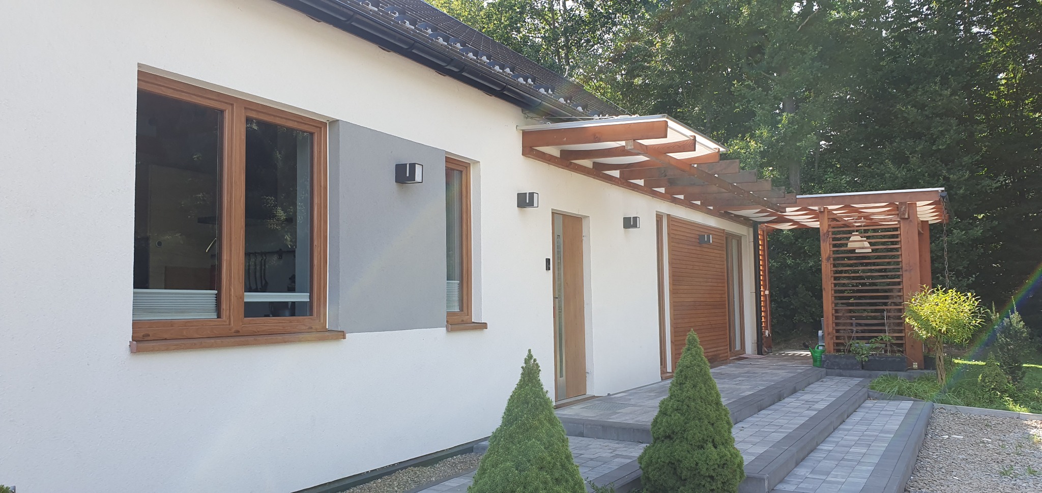 Nowoczesny dom szkieletowy z drewnianą pergolą i oknami w drewnianych ramach, widok z zewnątrz na elewację z dekoracyjnymi lampami i dwoma stożkowymi krzewami na pierwszym planie.