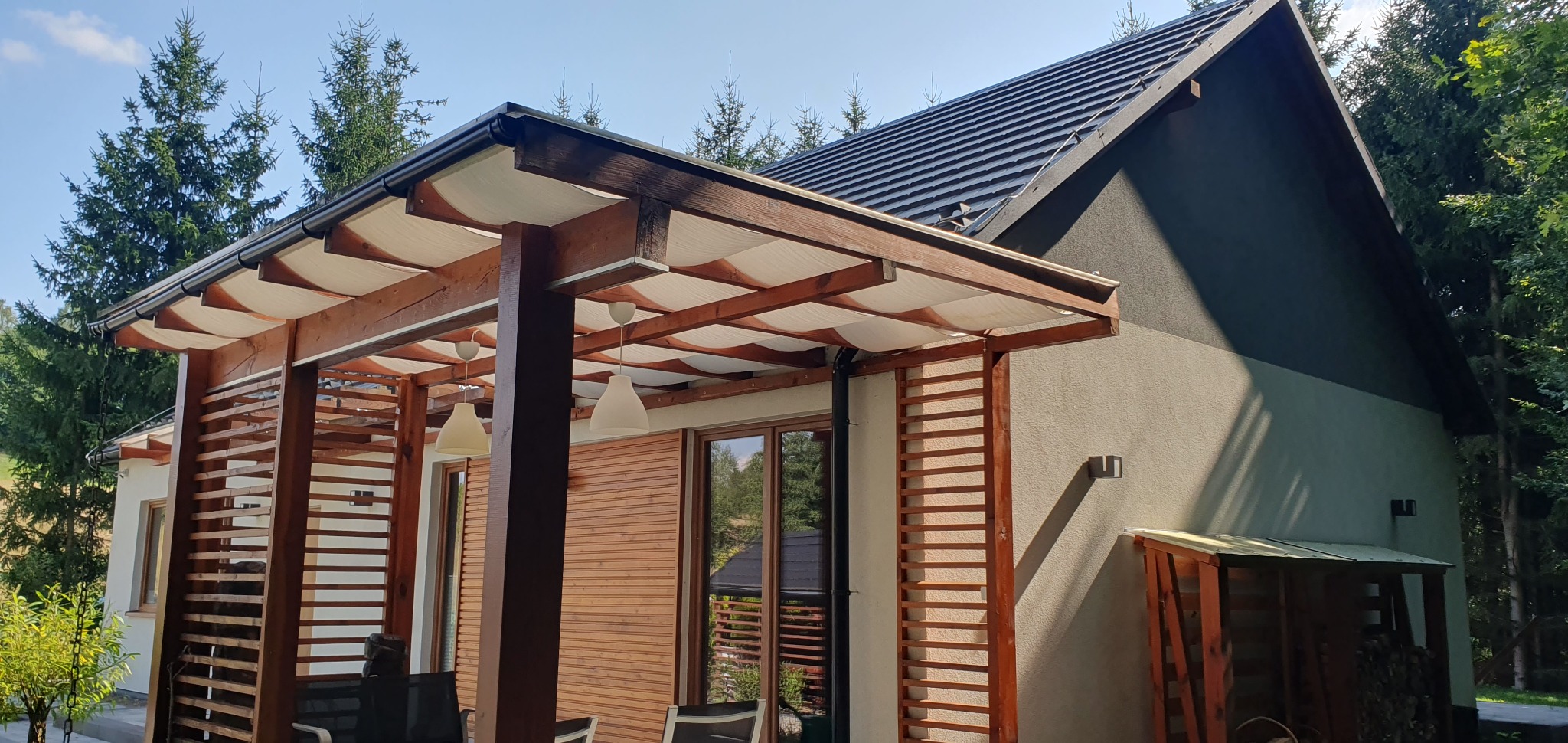 Nowoczesny dom z drewnianą pergolą z białym zadaszeniem, oświetloną wiszącymi lampami, na tle zieleni.