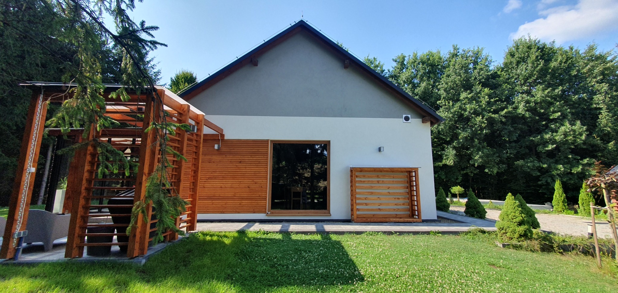 Nowoczesny dom z drewnianą elewacją i pergolą, otoczony zielonym trawnikiem i drzewami.