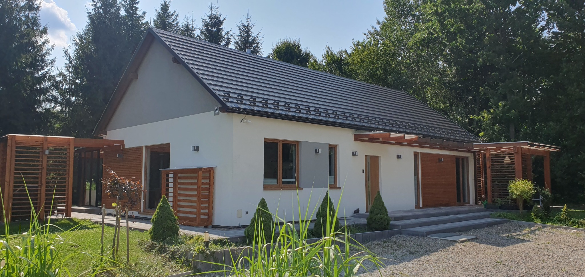 Nowoczesny parterowy dom z szarą dachówką i drewnianymi elementami dekoracyjnymi, otoczony zielenią i drzewami.