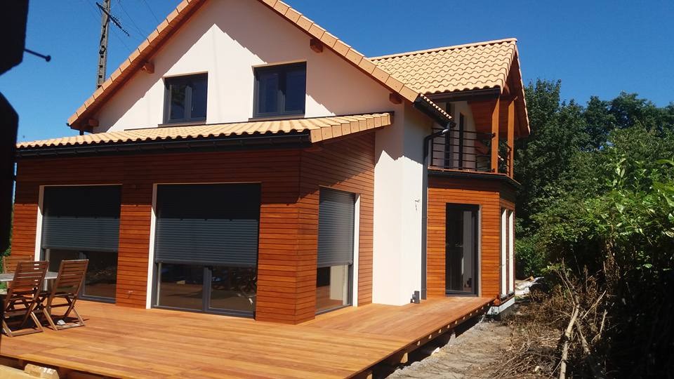 Nowoczesny dom z drewnianą elewacją i tarasem, rolety zewnętrzne częściowo zasłonięte, dwukondygnacyjny z balkonem, jasna elewacja górnej części budynku, dachówka ceramiczna.