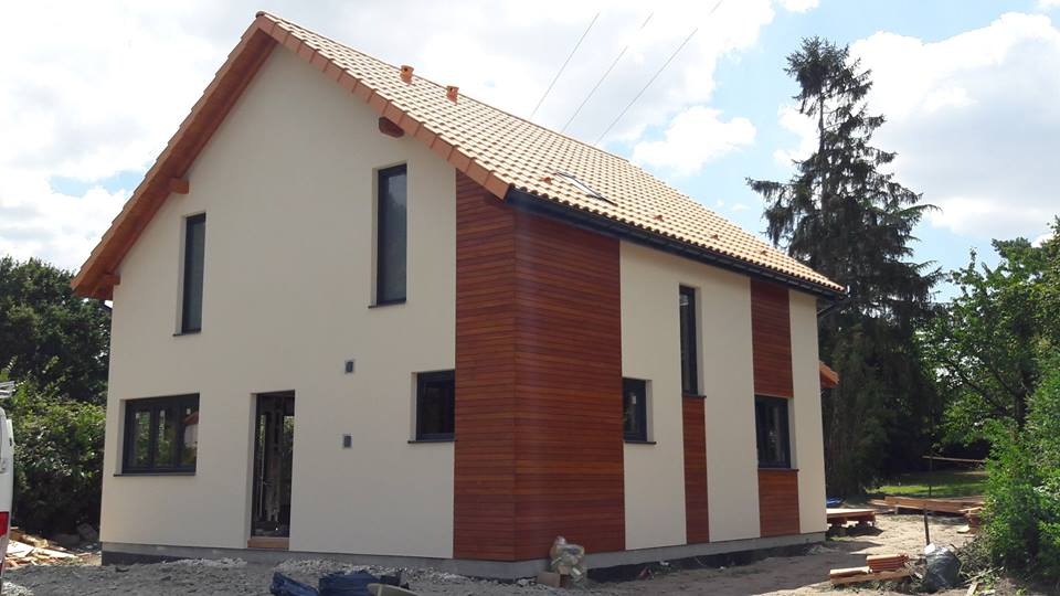 Dwukondygnacyjny budynek z jasną elewacją z drewnianymi akcentami, pomarańczowa dachówka, czarne ramy okienne, widok z zewnątrz w słoneczny dzień.