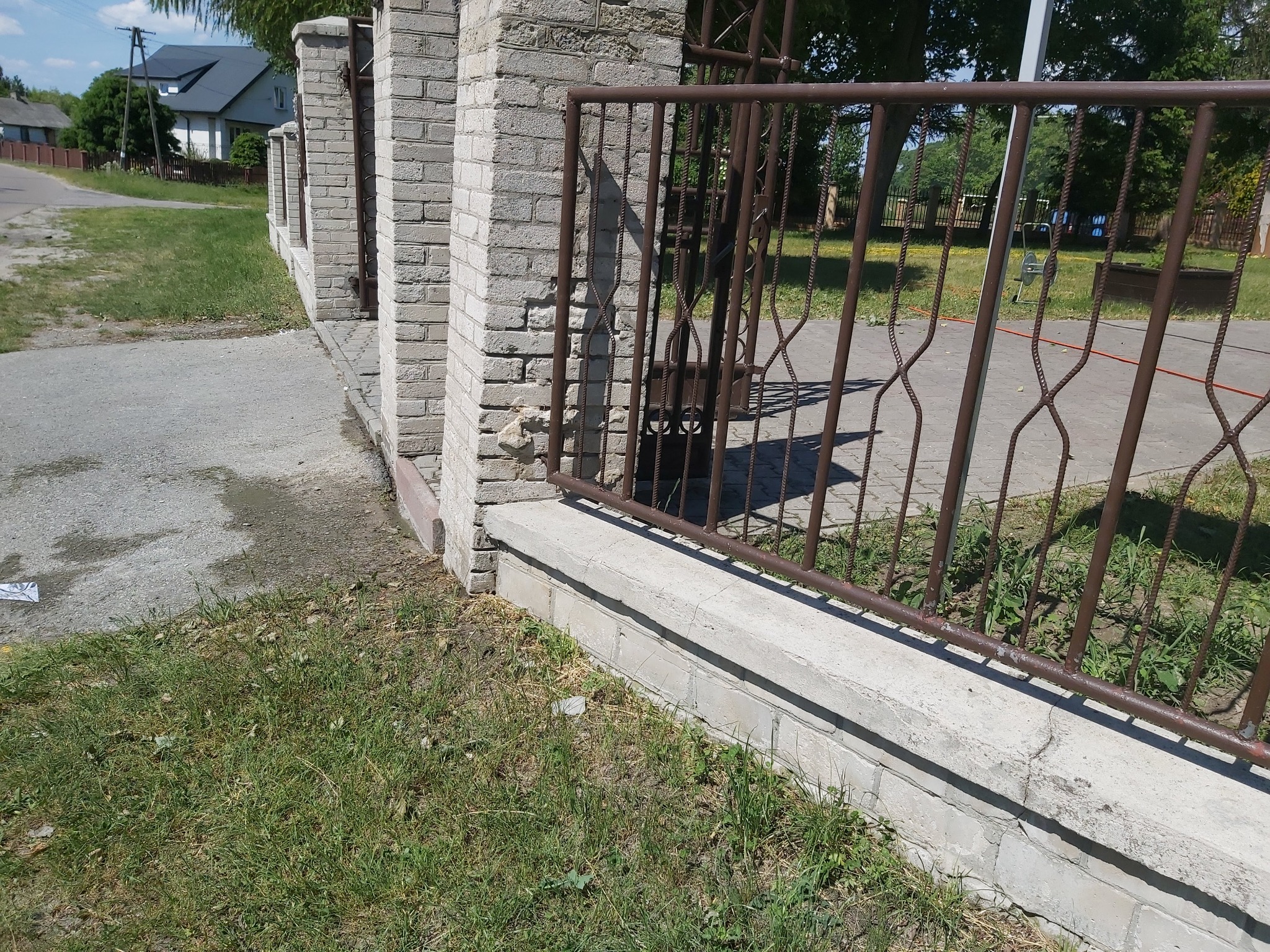 Fragment murowanego ogrodzenia z metalową, brązową bramą, widoczne uszkodzenia muru i nierówności terenu.