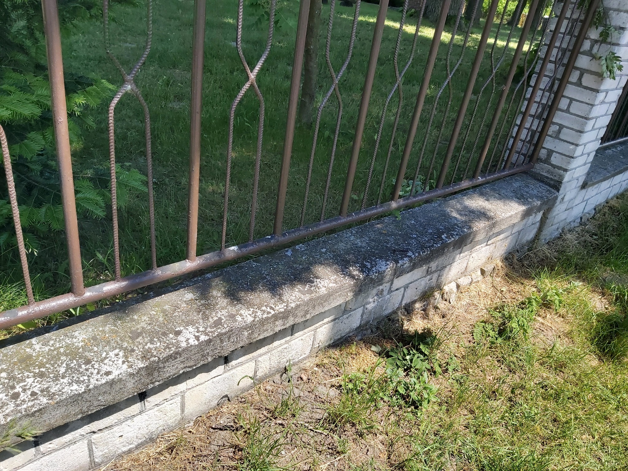 Metalowe ogrodzenie z dekoracyjnym wzorem, osadzone na betonowym murku z elementami cegły, otoczone trawą i zielenią.