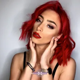 Portret kobiety z intensywnym makijażem oczu i ust, czerwone włosy w nieładzie, dłonie dotykają szyi, widoczny zegarek i bransoletka.