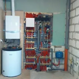 Kotłownia w trakcie montażu: piec gazowy kondensacyjny, zasobnik ciepłej wody i rozdzielacz ogrzewania podłogowego z licznymi czerwonymi rurami na tle surowych ścian z betonu komórkowego i płyt...