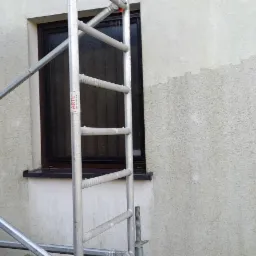 Fragment elewacji budynku z widocznym śladem mycia, rusztowaniem i klimatyzatorem z zabrudzeniami, okno z ciemną ramą.