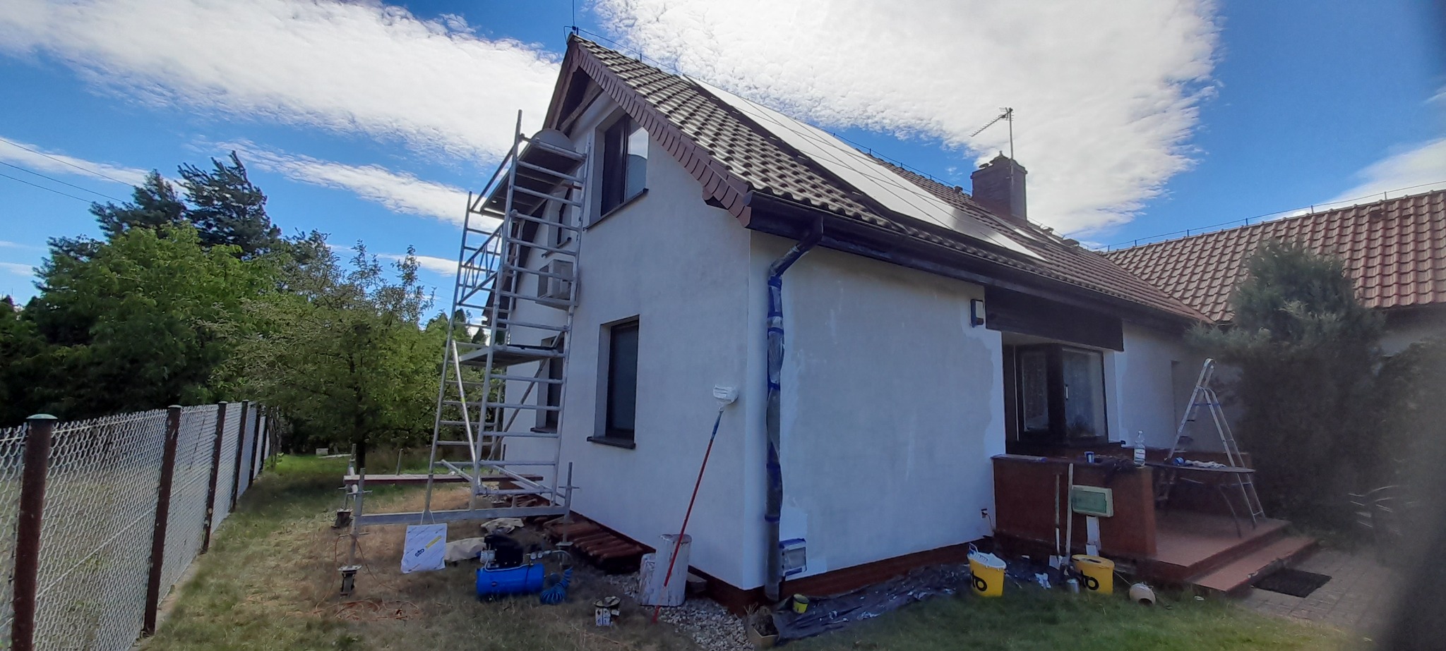 Dom jednorodzinny z częściowo pomalowaną elewacją na biało, widoczne rusztowanie, kompresor, wiaderka z farbą, zabezpieczone rynny, w tle błękitne niebo z chmurami i zieleń drzew