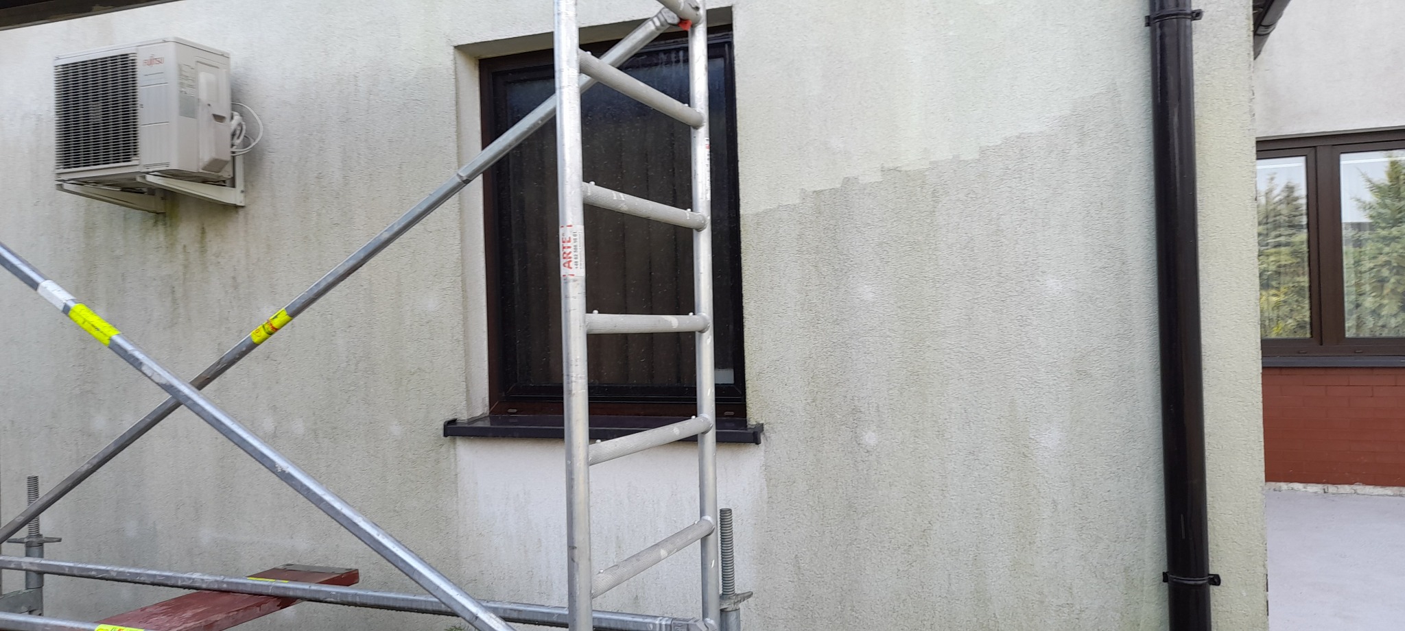 Fragment elewacji budynku z widocznym śladem mycia, rusztowaniem i klimatyzatorem z zabrudzeniami, okno z ciemną ramą.